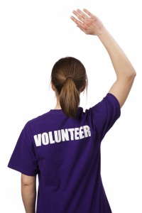 A young woman wearing a volunteer t-shirt stands against a pure white background with back to the camera. Her hand is raised, ready to help.